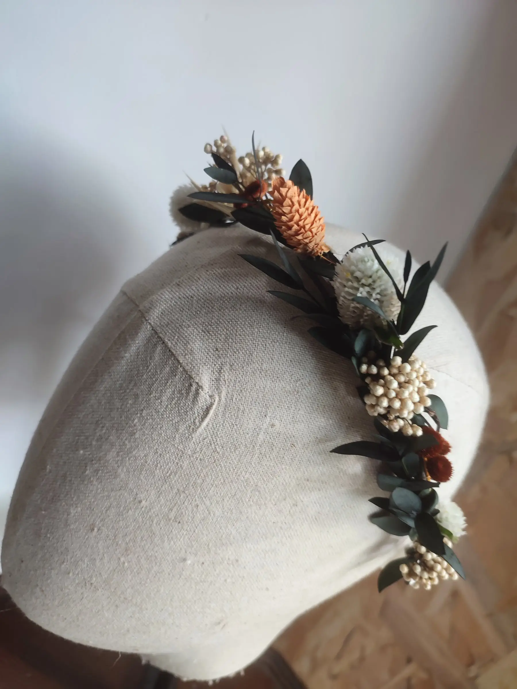 Couronne de fleurs séchées aux tons corail, crème et eucalyptus, posée sur une tête de modiste.